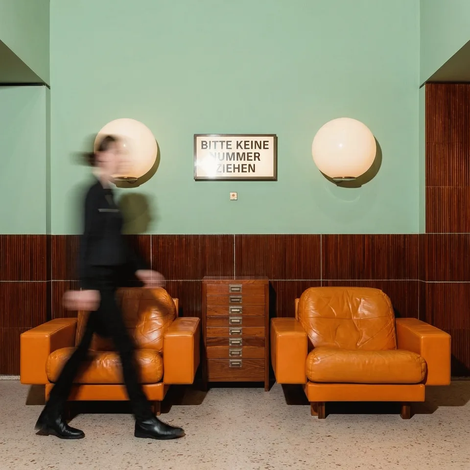 Amt Mitte, Markenraum: Lobby mit grüner Wand, zwei orangefarbenen Ledersesseln und einer vorbeigehenden Person